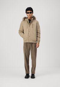 Bežna zip-up jakna s kapuco obloženo z mehkimi dlačicami, v kombinaciji s svetlo rjavimi kord pantami in črnimi čevlji. Model nosi sončna očala in stoji pred belo ozadje.