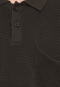 Tumši brūns polo kreklis ar teksturētu zigzaga rakstu, divreiz aizpogojams, ar apkakli. Mīksts trikotāžas audums ar vieglu spīdumu.