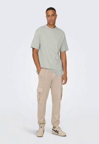 Camiseta verde claro de cuello redondo, combinada con pantalones cargo beige que cuentan con bolsillos laterales y un corte relajado. Zapatillas deportivas completan el atuendo.
