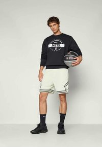 Felpa Nike nera con logo "NETS", abbinata a pantaloni corti da atletica bianchi con accenti neri, in piedi con un pallone da basket.