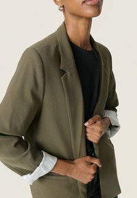 Blazer verde măsliniu cu rever în formă de notă, dintr-un material ușor, cu mâneci suflecate ce au dungi albe. Mâinile împreună la piept, dezvăluind o cămașă neagră dedesubt.