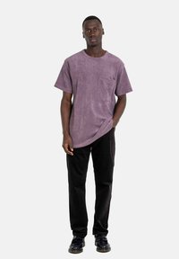 Lila Kurzarm-T-Shirt mit einer Brusttasche, aus weichem Stoff gefertigt. Kombiniert mit schwarzen Hosen und dunklen Schuhen, mit rundem Ausschnitt.