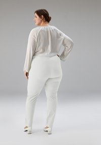 Blouse blanche en satin avec un col froncé et des manches longues, associée à un pantalon blanc ajusté et des chaussures à talons blocs blanches. Vue de dos.