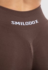 Bruine fitnesslegging met een geribbelde textuur en een hoge tailleband. De merknaam "SMILODOX" is prominent in het wit weergegeven aan de bovenkant.