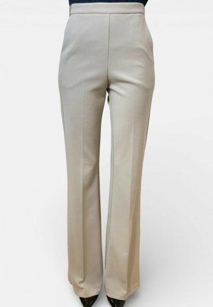 Pantaloni beige su misura a gamba dritta con vita alta, piega frontale e scarpe nere appuntite su uno sfondo semplice.