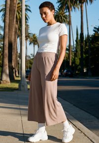 Young person with short hair wearing white T-shirt, beige wide-leg pants, white socks, and sneakers standing on sidewalk with palm trees behind.