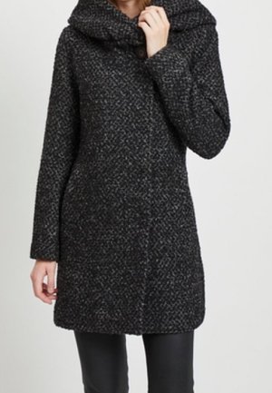 Femme portant un manteau en laine texturée gris foncé avec un col haut et large, des manches longues et un ourlet droit sur un pantalon noir.