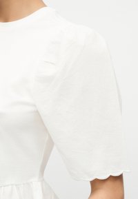 Λευκή κοντομάνικη μπλούζα με φουσκωτά μανίκια, με κυματιστό τελείωμα και ελαφριά ρυτίδωση στους ώμους σε ανοιχτόχρωμο δέρμα.