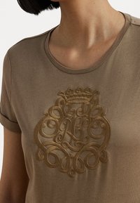 Camiseta de algodón beige con un diseño circular en relieve bordado en oro, que presenta intrincados remolinos y un motivo de corona en el centro.