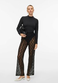 Črne čipkaste hlače z visokim pasom, okrašene z cvetličnim vzorcem in prosojno teksturo, v kombinaciji z dolgimi črnimi rokavi in belimi petkami.