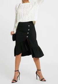 Svart, knä-lång kjol med volangkant, med en slits framtill och fem metallknappar. Bärs med en vit kabelstickad tröja och svarta klackar.