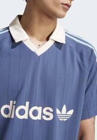 Blå sportströja med V-hals och vit krage. Har vertikala ränder och vit branding fram. Accentuerad med tre ränder på axlarna.