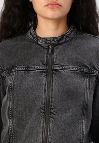Chaqueta de denim gris con un cuello alto, cremallera visible y cierre a presión. Presenta costuras horizontales y dos bolsillos en el pecho. La textura parece ligeramente desgastada.
