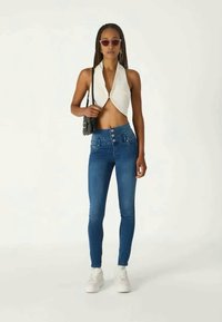 Top corto blanco con cuello halter, vaqueros ajustados de tiro alto azul con tres botones frontales, combinado con zapatillas blancas y gafas de sol oscuras.