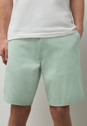 Mann trägt hellgrüne knielange Shorts und ein weißes T-Shirt mit den Händen in den Taschen.