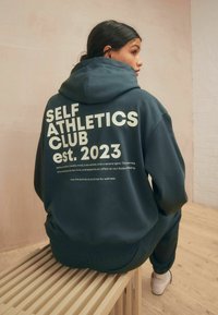 Tamsiai žalia džemperis su balta užrašu "SELF ATHLETICS CLUB est. 2023." Su raišteliu reguliuojamu gaubtu ir gumbuotomis rankovėmis. Dėvimi su atitinkančiomis kelnėmis.