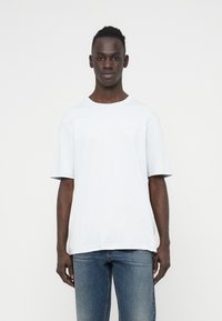 Λευκός κοντομάνικος T-shirt σε χαλαρή εφαρμογή, κατασκευασμένο από μαλακό ύφασμα με στρογγυλή λαιμόκοψη και διακριτική λεπτομέρεια λογότυπου στο στήθος. Συνδυασμένο με μπλε τζιν.