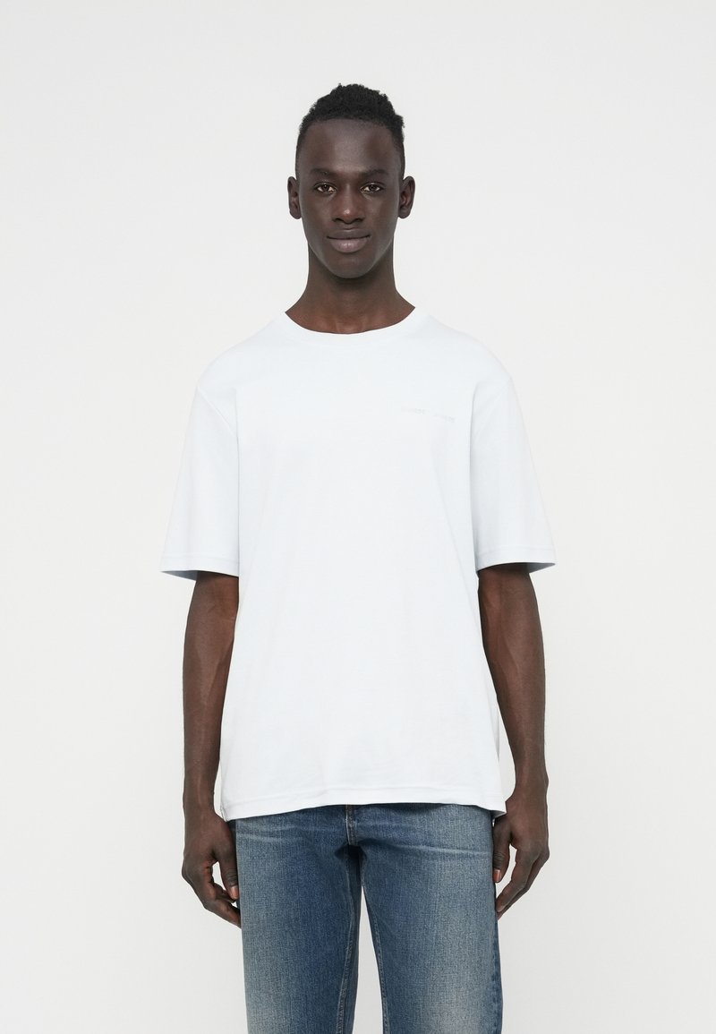 Λευκός κοντομάνικος T-shirt σε χαλαρή εφαρμογή, κατασκευασμένο από μαλακό ύφασμα με στρογγυλή λαιμόκοψη και διακριτική λεπτομέρεια λογότυπου στο στήθος. Συνδυασμένο με μπλε τζιν.