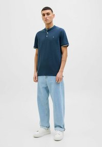 Marineblaues Poloshirt mit Kragen und drei Knöpfen, ausgestattet mit grün gestreiften Ärmeln, kombiniert mit hellblauen, weit geschnittenen Jeans und weißen Sneakers.
