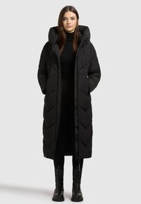 Sort lang pufferjakke med et chevron-mønster, oversized hætte, frontlynlås og justerbare snore. Båret over sort tøj.