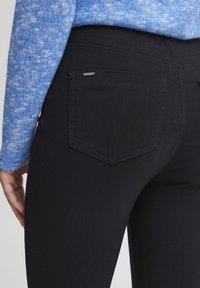 Črne jeans hlače s prilegajočim dizajnom, ki imajo zadnji žep s šivi in majhno oznako blagovne znamke. Vidna je tudi modra pulover s teksturo.