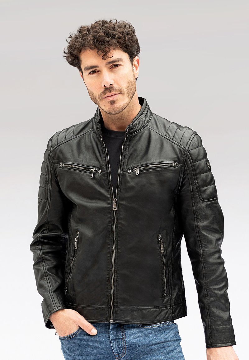 Mann mit lockigem Haar, der eine schwarze Lederjacke mit Reißverschluss und Reißverschlusstaschen sowie blaue Jeans trägt, Hände in den Taschen, neutraler Hintergrund.