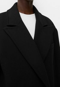 Svart ullblandad rock med slagkrage, en ficka och texturerad tyg. Vit crew neck-tröja synlig under.