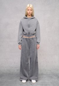 Grijze cropped hoodie met een ritssluiting en capuchon, gedragen met losse grijze sweatpants. Soepele textuur, casual pasvorm, met elastische tailleband. Witte sneakers.