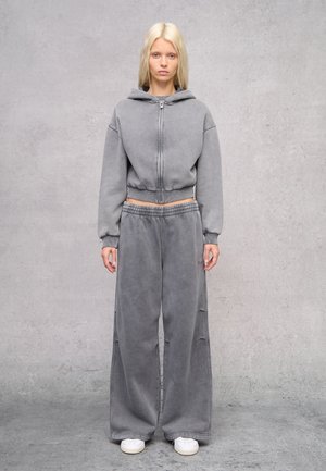 Felpa grigia corta con zip e cappuccio, abbinata a pantaloni della tuta grigi larghi. Tessuto liscio, vestibilità casual, con vita elastica. Sneakers bianche.