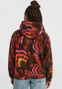Bruine fleecetrui met grote kleurrijke bloemenpatronen in roze, oranje en geel, met een ontspannen pasvorm en een capuchon met trekkoord.