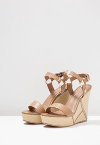 Sandales compensées beiges avec un design strappy, tige en cuir lisse et texture tissée sur la plateforme. Présente des accents en métal doré.