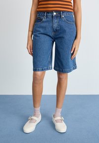 Demin bermudashorts i mellomblå farge med en avslappet passform, synlige sømmer og frontlommer. Kombinert med lyse sko og sokker.
