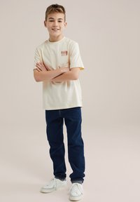 T-shirt z krótkim rękawem w kolorze kremowym z logo "BLUE RIDGE", zestawiony z ciemnoniebieskimi dżinsami i białymi trampkami.