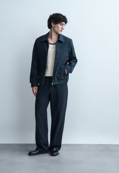 Νεαρός άνδρας με σγουρά μαλλιά που φοράει σκούρο σακάκι με σχέδια, μπεζ πουκάμισο, μαύρο παντελόνι φαρδιάς γραμμής και μαύρα παπούτσια, στέκεται μπροστά σε έναν απλό τοίχο.