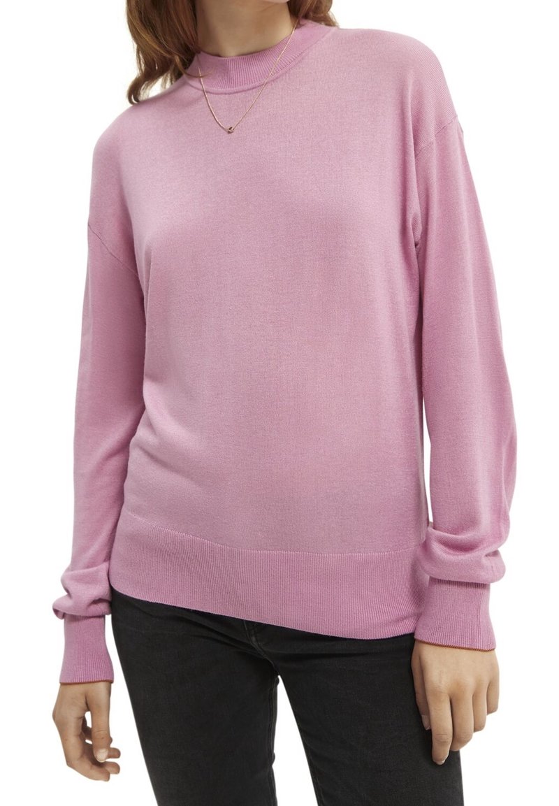 Femme portant un pull en maille rose à manches longues et un pantalon noir, avec un délicat collier en or visible au niveau du col.