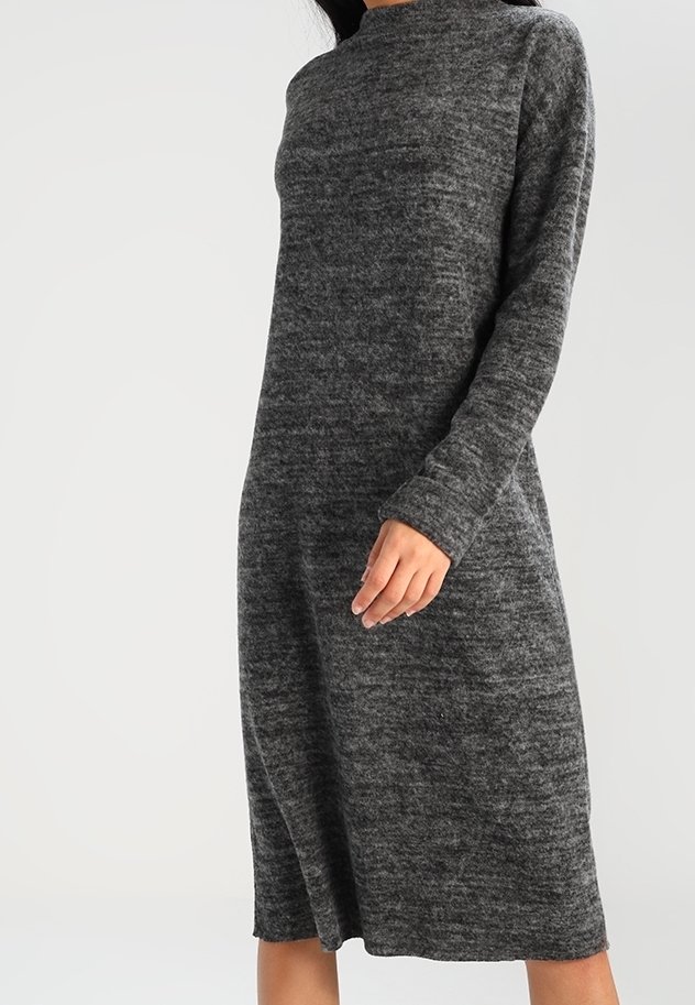 Femme portant une robe en maille gris foncé à manches longues, longueur genou, avec un col montant, sur un fond clair uni.
