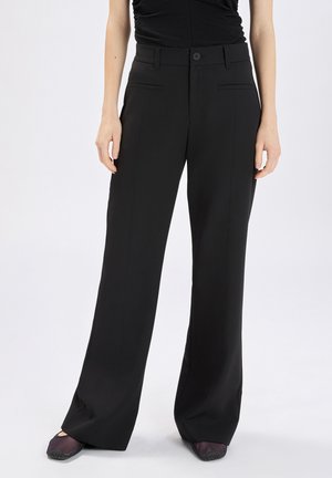 Donna che indossa pantaloni neri a gamba larga e vita alta con tasche frontali e chiusura a bottoni, abbinati a scarpe scure, in piedi su sfondo bianco.