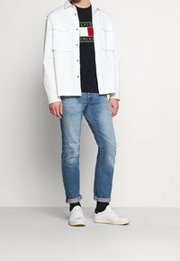 Muž oblečený v bílém zapínacím saku, černém tričku s grafikou, světlých modrých džínách s vyhrnutými nohavicemi, černých ponožkách a bílých teniskách, stojící na jednobarevném pozadí.