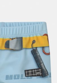 Calças curtas azul claro com um cinto amarelo, apresentando um gráfico de uma lanterna preta e um rótulo preto com um design de grelha branca, com a inscrição "Molo."