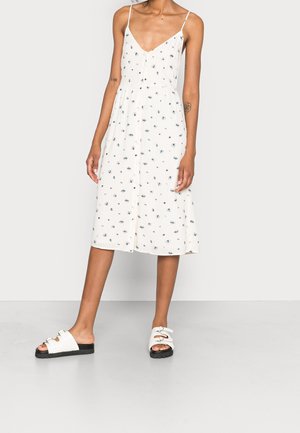 Woman wearing a white midi dress with thin straps and small blue flower patterns, paired with white slip-on sandals with black soles.