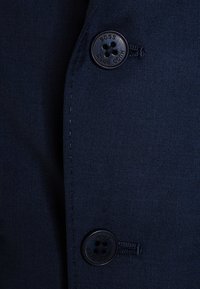 Zbliżenie na granatowy materiał z dwukluczowym zapięciem, o teksturowanym wykończeniu i czarnym guzikiem z wytłoczonym napisem "Hugo Boss".