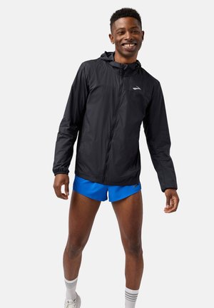 Leichte schwarze Laufjacke mit Kapuze, langen Ärmeln und reflektierendem Logo; kombiniert mit leuchtend blauen Shorts und weißen Socken.
