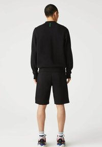 Sudadera negra y pantalones cortos, material de forro polar, corte relajado. Etiqueta verde notable en la parte posterior del cuello. Usado con zapatillas deportivas coloridas.
