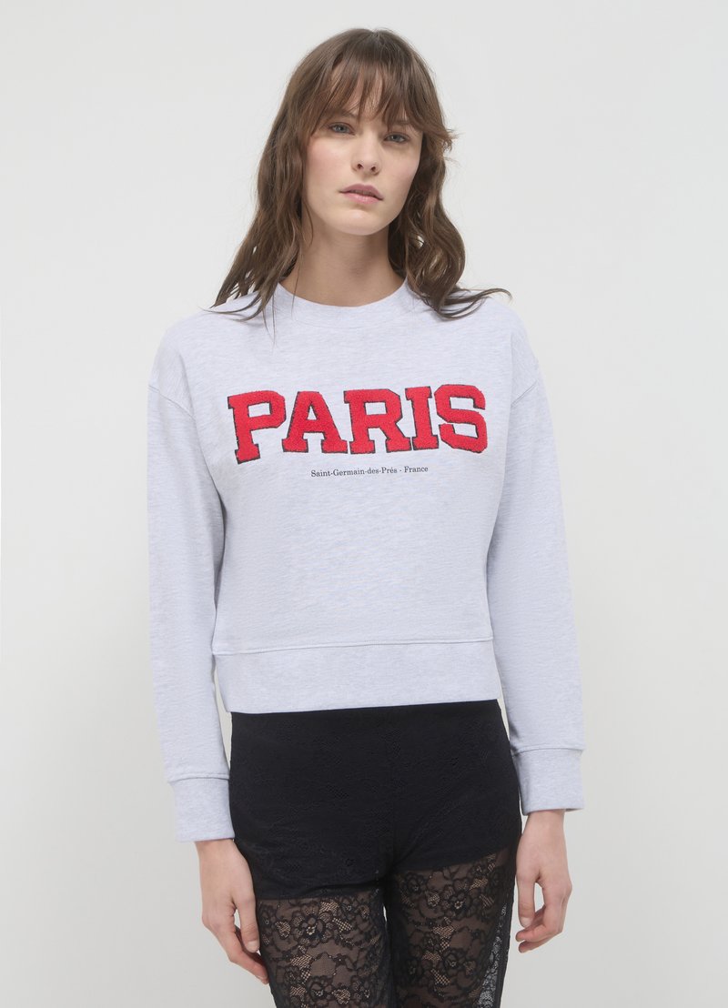 Sweatshirt gris clair avec un grand lettrage rouge "PARIS" et un texte plus petit en dessous. Design cropped avec des manches longues et des bords côtes.