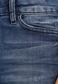 Tela de denim azul oscuro con una textura desgastada, mostrando una costura de bolsillo y detalles de puntadas. El material es suave y duradero.