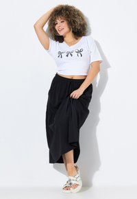 Weißes, kurz geschnittenes T-Shirt mit schwarzen Schleifengrafiken, kombiniert mit einem schwarzen Maxirock. Das Modell trägt silberne Sandalen und zeigt einen entspannten Schnitt und Stil.