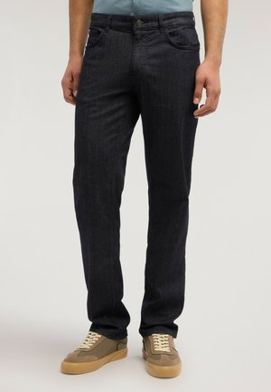 Hombre con jeans oscuros de corte recto y zapatillas beige, de pie contra un fondo neutro.