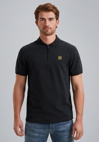 Černé polo tričko s límcem na zip a krátkým rukávem. Má malou žlutou nášivku na hrudi a texturovanou tkaninu. Nošeno s dením džínami.