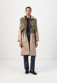 Manteau léger beige avec taille à cordon, associé à un gilet rembourré vert. Comprend des poches avant et un design avec col. Les chaussures sont noires.