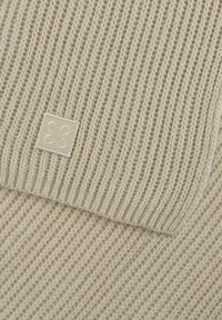 Écharpe en tricot tissé de couleur crème avec une texture côtelée. Présente une étiquette en tissu carré avec un design de fleur stylisé à un coin.